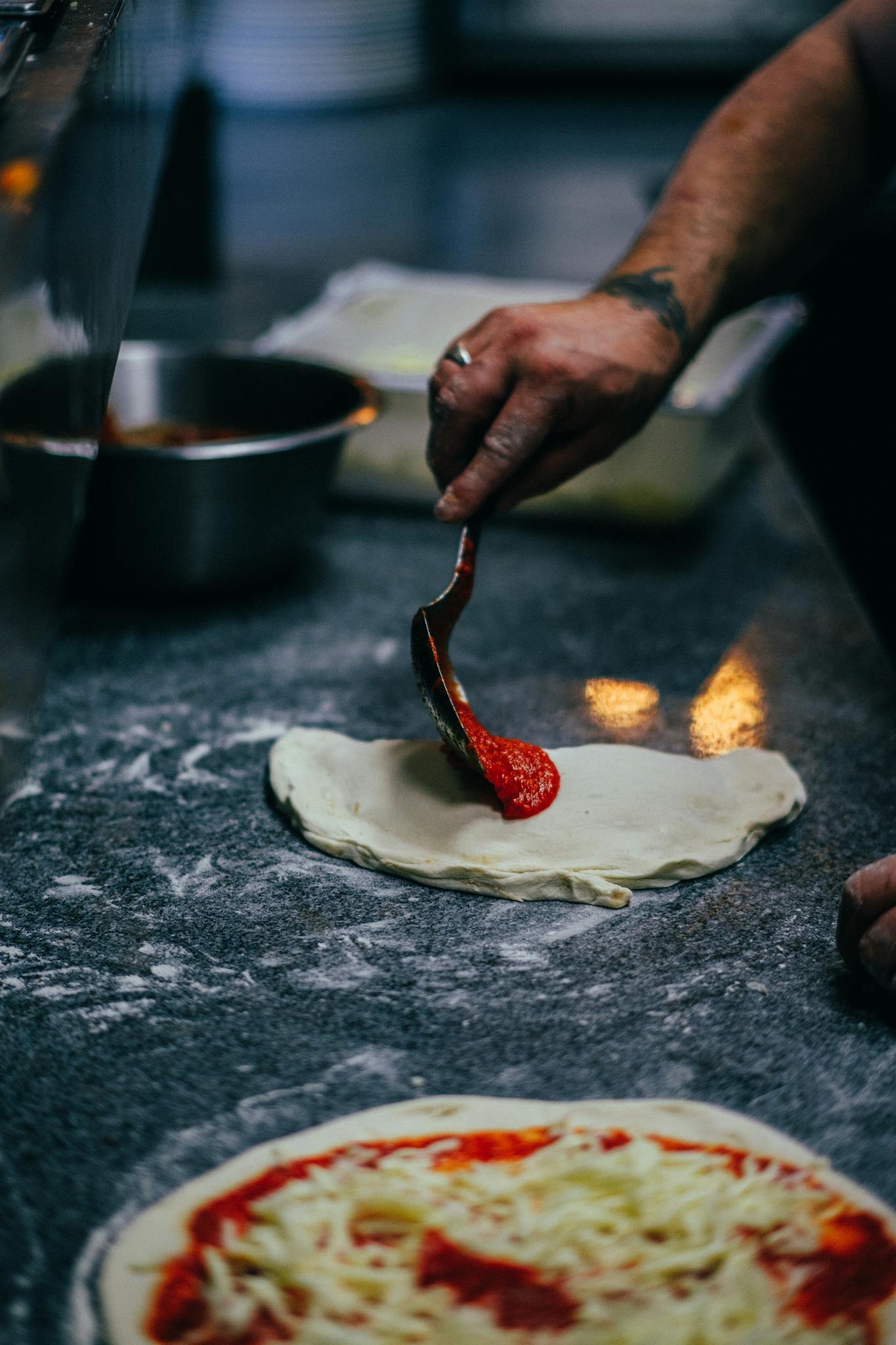 A close-up of homemade pizza dough being topped with tomato sauce by hand in a cozy kitchen setting.