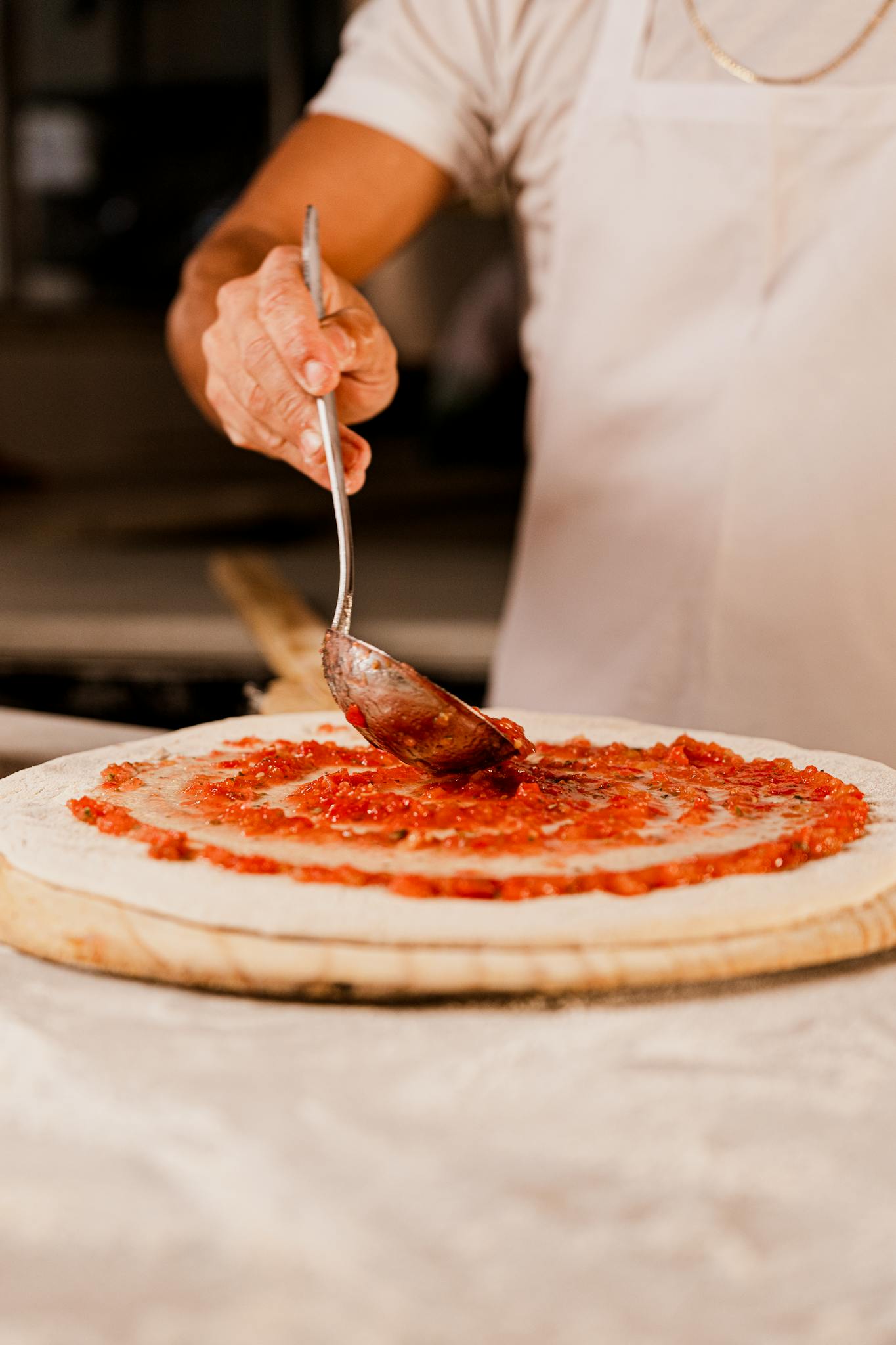 Chef spreading tomato sauce on pizza dough in a kitchen setting with a spoon.